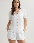 A woman with long brown hair wears a Bella Dahl kurzarmbluse with Hemdkragen and chest pocket, paired with matching white shorts. She stands confidently, hands in pockets, against a plain, light-colored background.