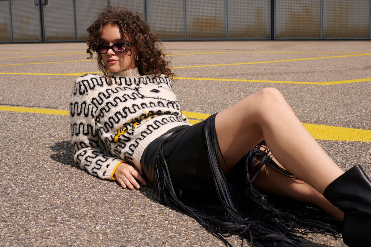Frau trägt schwarzen Lederrock und weissen Snoopy-Wollpullover mit handgestrickter Intarsie, liegt entspannt auf Asphalt mit gelben Linien, modern und stilvoll inszeniert.