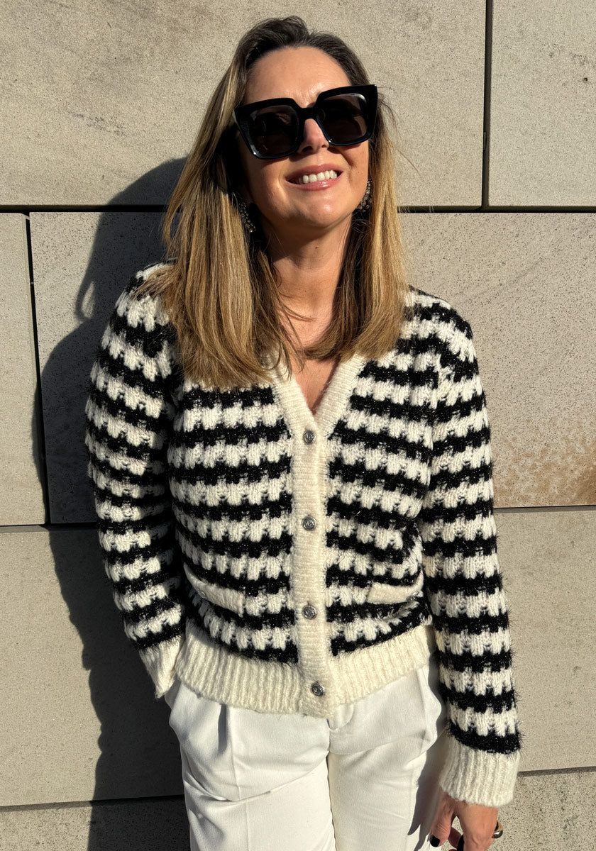 A woman wearing black sunglasses, a black-and-white striped button-up sweater, and white pants stands smiling against a beige stone wall in bright sunlight.