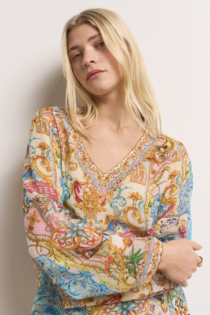 Eine junge Frau mit langen blonden Haaren posiert vor einer weißen Wand und trägt eine bunte, gemusterte Bluse mit V-Ausschnitt und floralen und verschnörkelten Mustern in Rosa-, Gold-, Blau- und Grüntönen.