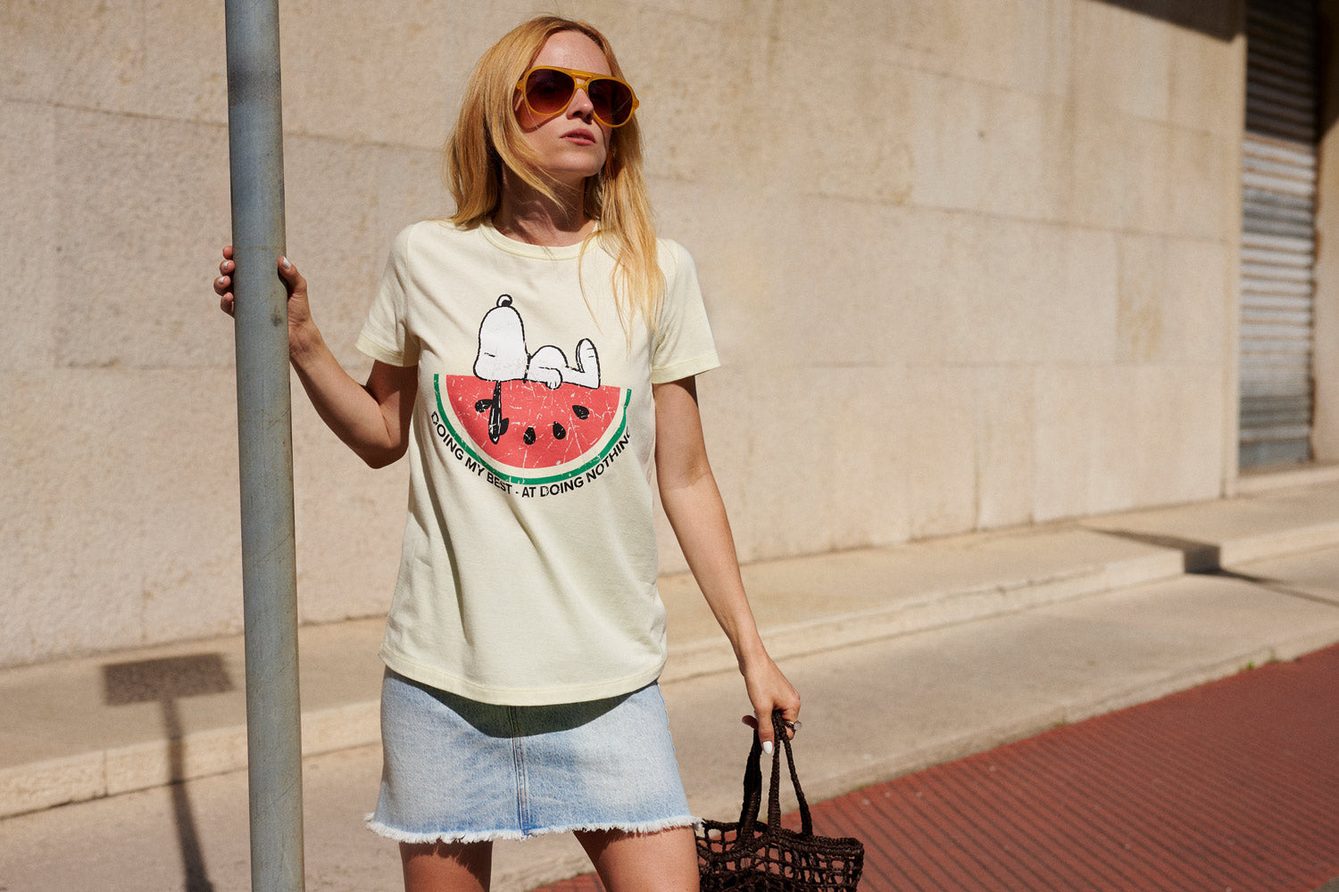 Eine Frau mit Sonnenbrille trägt ein T-Shirt mit Snoopy-Motiv und einen Jeansrock; sie steht draußen neben einem Pfosten und hält eine schwarze Tasche in der Hand, im Hintergrund sind eine beige Wand und ein roter Bürgersteig zu sehen.
