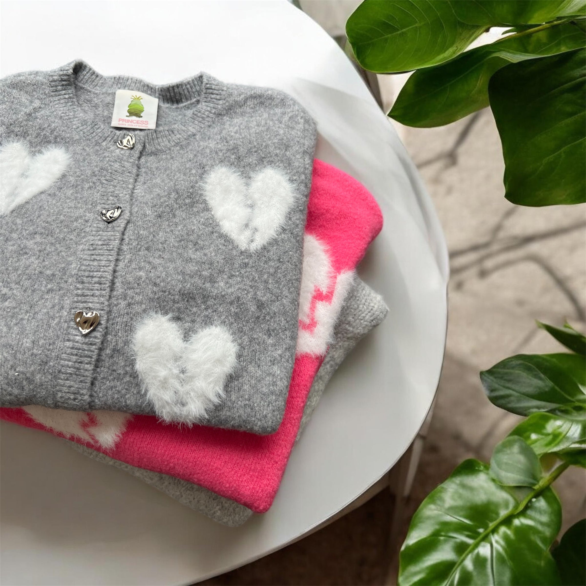 Eine Frau mit langen braunen Haaren trägt einen schwarzen Pullover mit einem Muster aus großen weißen gebrochenen Herzen. Sie blickt vor einem schlichten hellen Hintergrund nach vorne und hat einen neutralen Ausdruck.