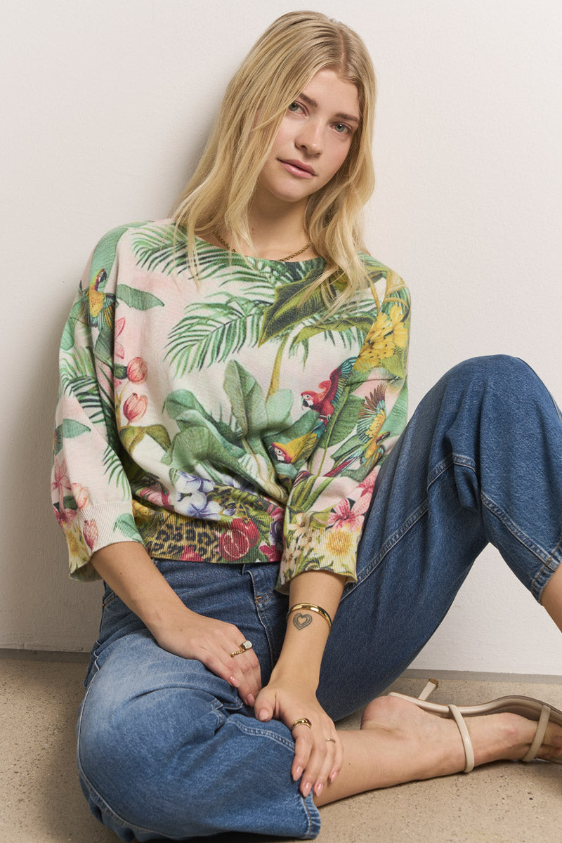 Eine junge Frau mit langen blonden Haaren, die einen bunten Pullover mit tropischen Motiven und blaue Jeans trägt, sitzt auf dem Boden vor einer weißen Wand, hat ein Knie hochgezogen und einen Arm auf ihr Knie gestützt.