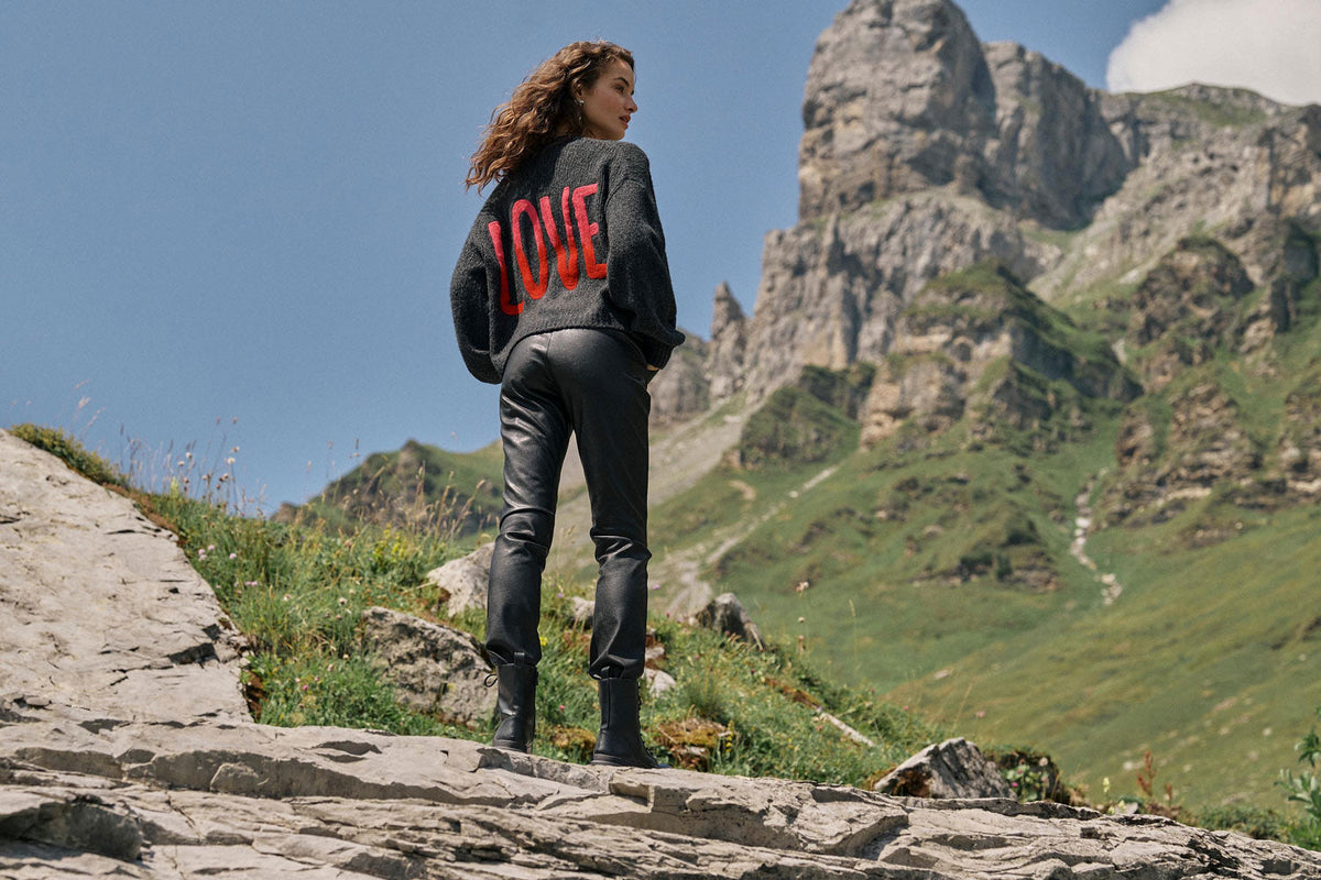 Eine Frau in schwarzer Hose und Stiefeln steht auf felsigem Gelände und blickt auf einen Berg. Auf dem Rücken ihres Pullovers steht in roten Buchstaben „LOVE“. Hinter ihr erheben sich grüne Hänge und schroffe Gipfel unter einem blauen Himmel mit Wolken.