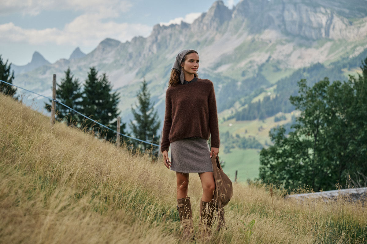 Eine Frau in einem braunen Pullover, einem grauen Rock und braunen Stiefeln steht auf einem grasbewachsenen Hang und hält eine braune Tasche in der Hand. Im Hintergrund sind Kiefern und hohe Berge unter einem teilweise bewölkten Himmel zu sehen.