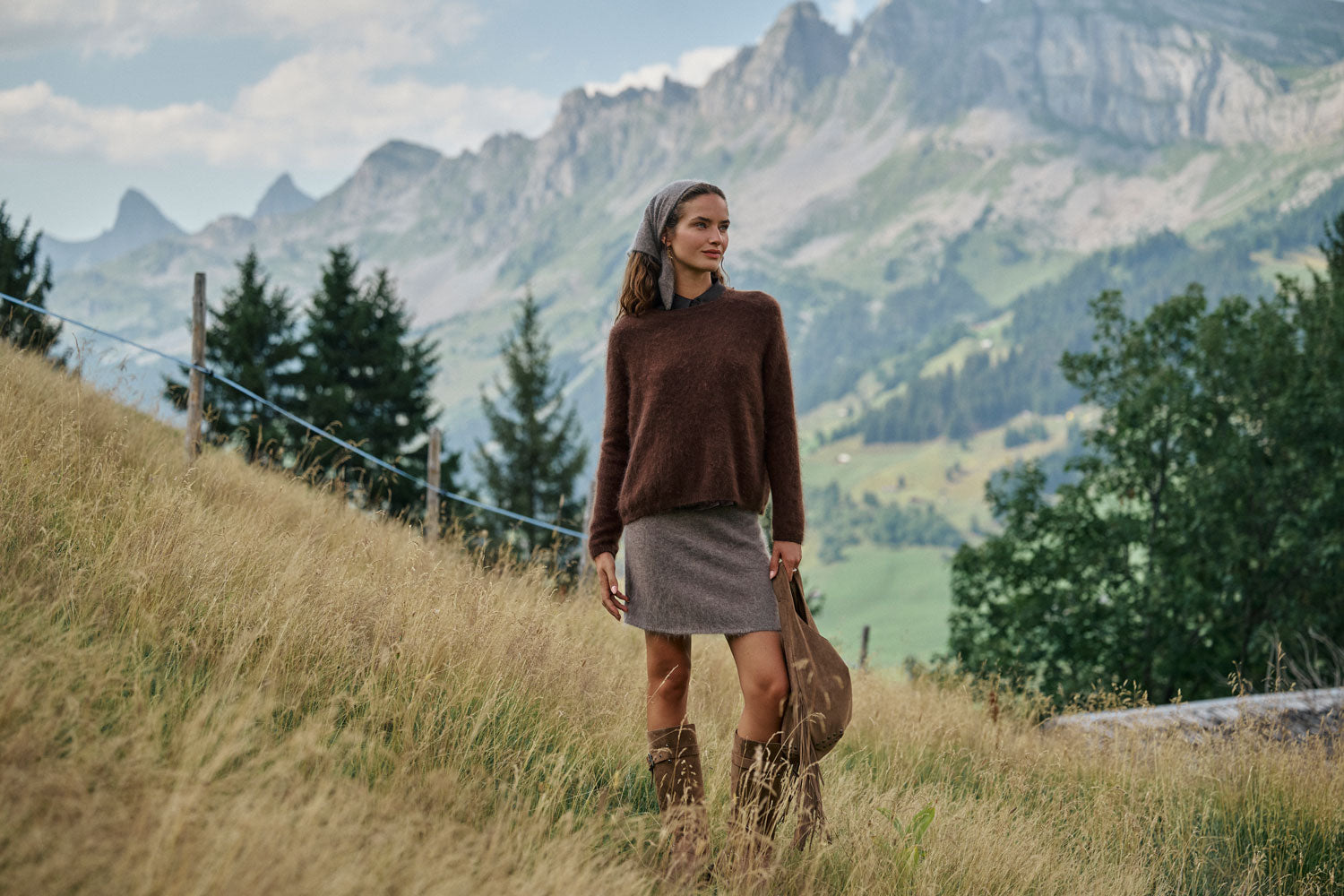 Eine Frau steht auf einem grasbewachsenen Hang, sie trägt einen braunen Pullover, einen grauen Rock und Stiefel und hält eine Tasche in der Hand. Im Hintergrund sind Berge, Bäume und ein bewölkter Himmel zu sehen.