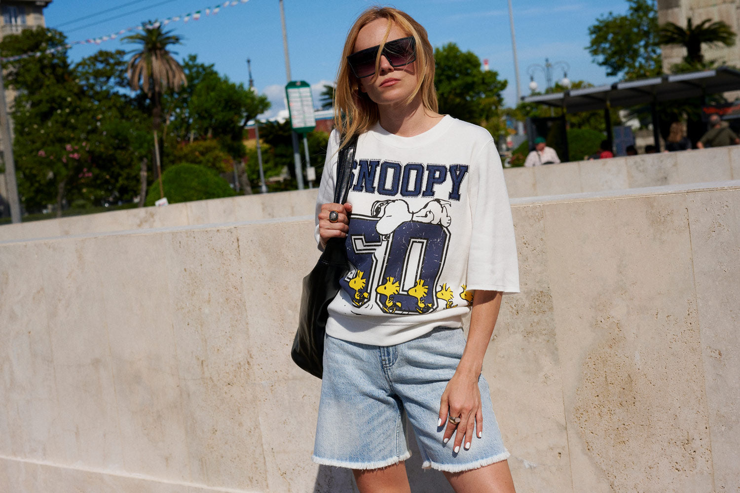 Eine Frau mit Sonnenbrille und einem weißen T-Shirt mit Snoopy-Motiv sowie Jeansshorts steht im Freien vor einer Steinmauer und hält eine schwarze Tragetasche in der Hand; im Hintergrund sind Bäume und ein blauer Himmel zu sehen.