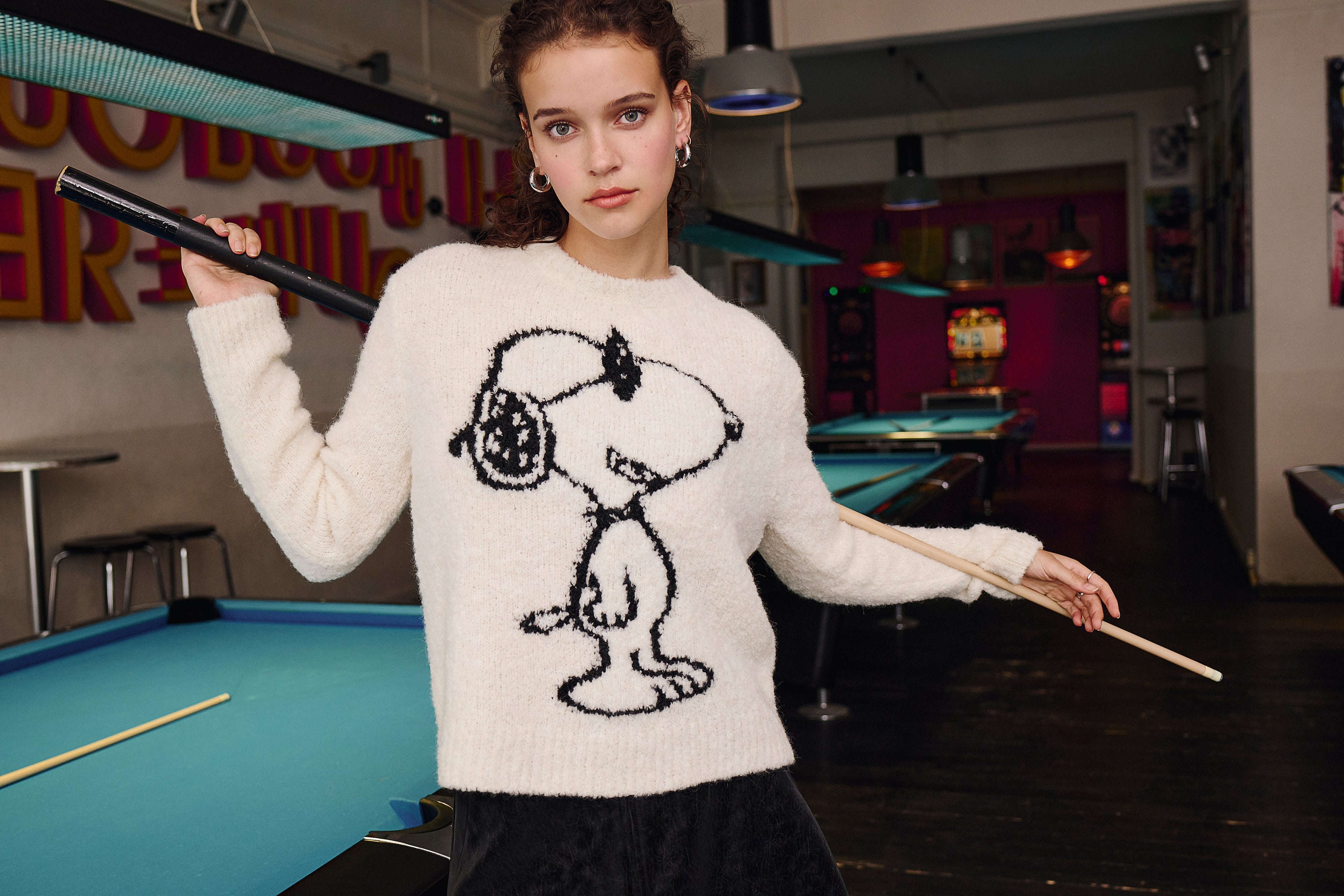 A young woman in a white Snoopy sweater stands in a pool hall, holding a pool cue and a pool stick, with blue pool tables and neon lights in the background.