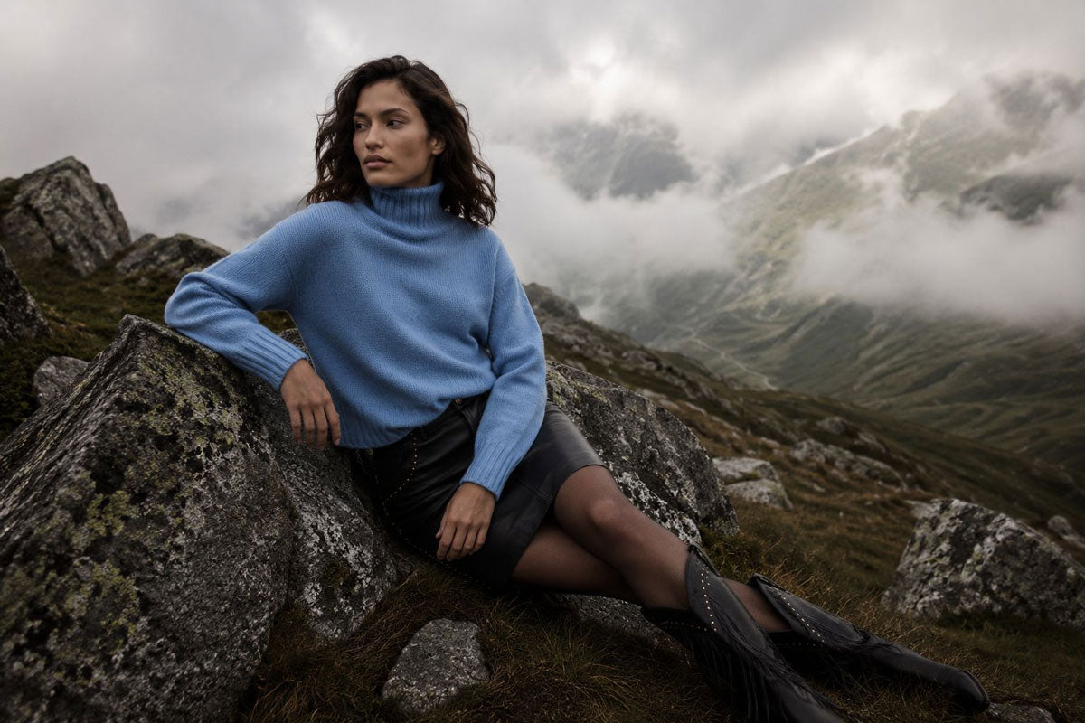 Eine Frau in einem blauen Rollkragenpullover und einem schwarzen Rock sitzt auf einem felsigen Berghang. Im Hintergrund zieht Nebel über schroffe Berggipfel hinweg.