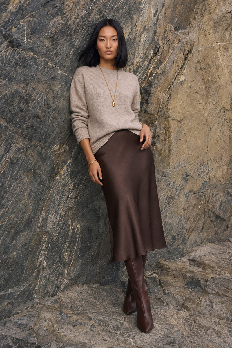 Eine Frau mit langen schwarzen Haaren trägt einen beigen Pullover, eine goldene Halskette, einen braunen Midirock aus Satin und braune kniehohe Stiefel und lehnt an einer strukturierten Steinmauer.