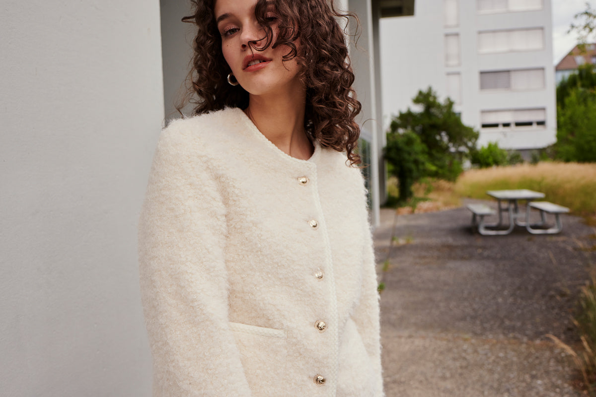 A woman with curly brown hair wears a textured, cream-colored button-up jacket, standing outdoors near a pale building with greenery, a picnic table, and an overgrown lawn in the background.
