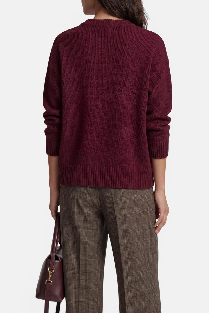 Eine Person, von hinten gesehen, die einen dunkelroten Kaschmirpullover mit Rundhalsausschnitt und eine braune Karohose trägt und eine dazu passende dunkelrote Handtasche an der Seite hält.
