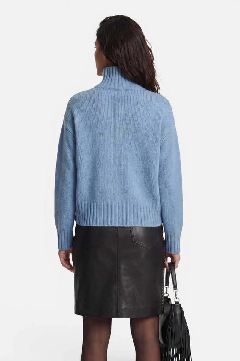 Eine Person, die von hinten zu sehen ist, trägt einen hellblauen Rollkragenpullover aus Kaschmir und einen schwarzen, knielangen Lederrock und hält eine schwarze Lederhandtasche mit Fransenverzierung in der Hand.