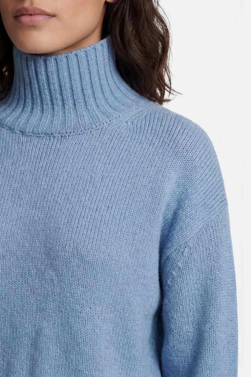 Eine Person trägt einen hellblauen, gerippten Kaschmir-Rollkragenpullover. Die Nahaufnahme konzentriert sich auf den hohen Kragen und die Struktur des Strickstoffs vor einem schlichten weißen Hintergrund.