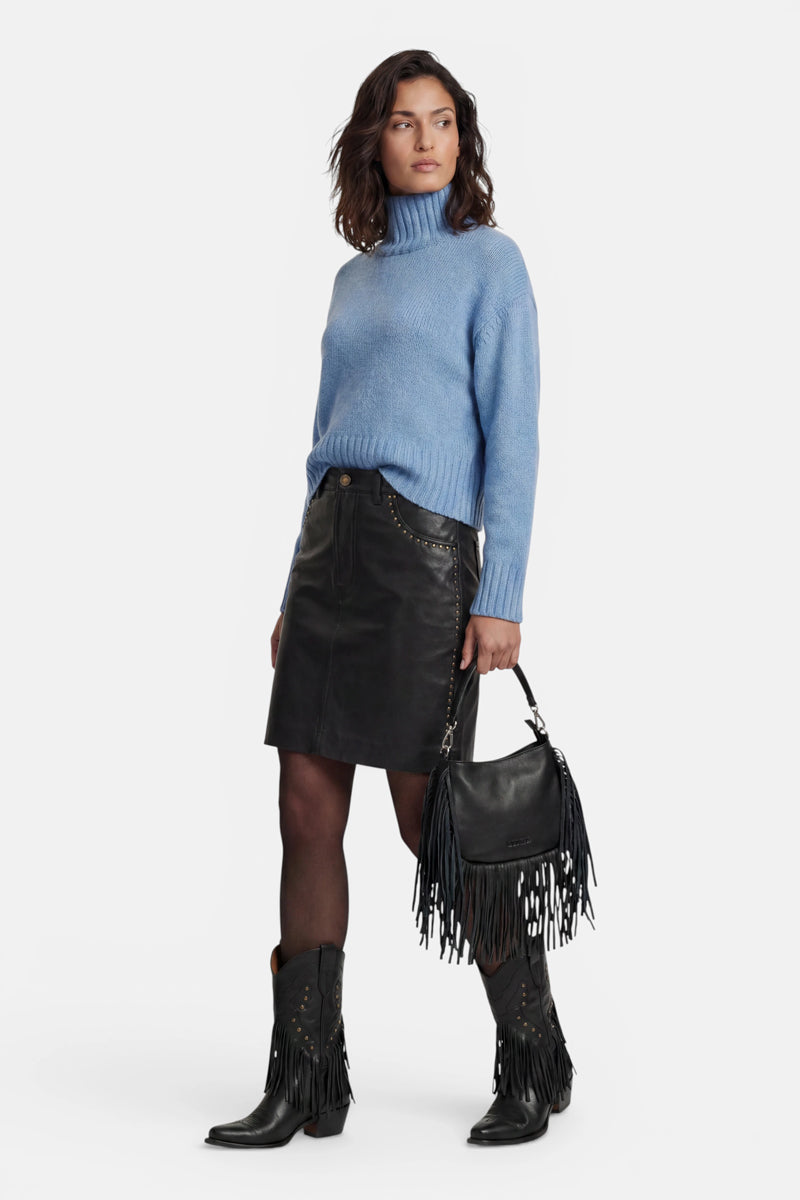 Eine Frau, die einen hellblauen Kaschmir-Rollkragenpullover, einen schwarzen Lederrock mit goldenen Nieten und schwarze Cowboystiefel trägt. Sie steht vor einem weißen Hintergrund und hält eine schwarze Lederhandtasche mit langen Fransen in der Hand.