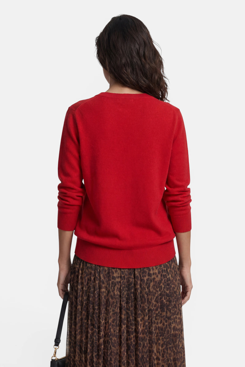 Eine Person mit dunklen Haaren, von hinten gesehen, trägt einen leuchtend roten Kaschmirpullover mit V-Ausschnitt. Dazu trägt sie einen braun-schwarzen Plisseerock mit Leopardenmuster und eine kleine schwarze Handtasche.