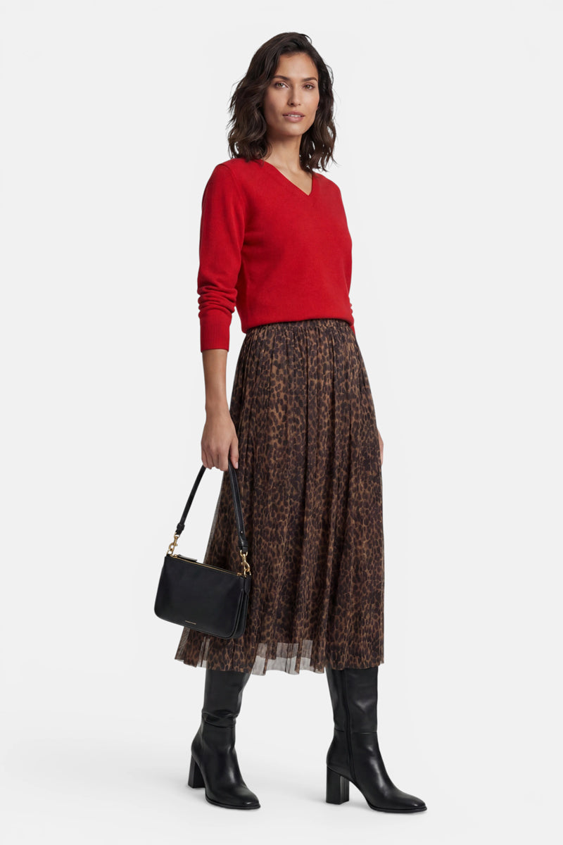 Eine Frau trägt einen roten Kaschmirpullover mit V-Ausschnitt und einen plissierten Midirock mit Leopardenmuster. Sie hat eine schwarze Umhängetasche dabei und trägt kniehohe schwarze Stiefel vor einem schlichten weißen Hintergrund.