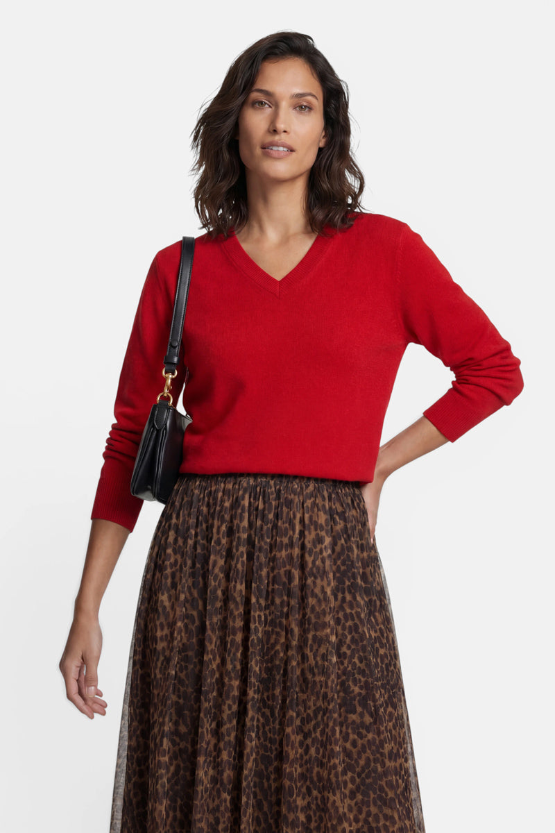 Eine Frau mit schulterlangem braunem Haar posiert vor einem schlichten weißen Hintergrund in einem leuchtend roten Kaschmirpullover mit V-Ausschnitt und einem braunen Midirock mit Leopardenmuster. Sie trägt eine kleine schwarze Umhängetasche mit einem goldenen Kettenriemen.