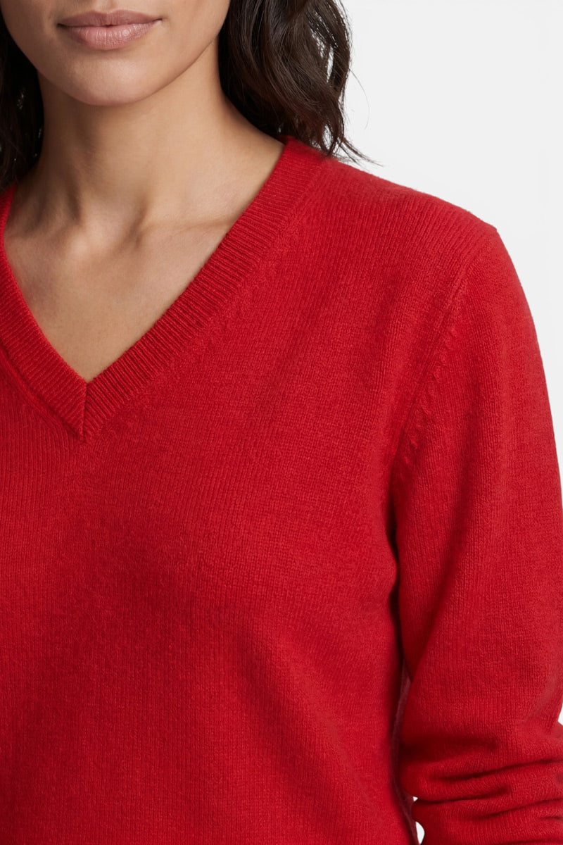 Eine Frau mit dunklem Haar trägt einen leuchtend roten Kaschmirpullover mit V-Ausschnitt. Die Nahaufnahme hebt die Struktur des Strickstoffs und den Ausschnitt vor einem schlichten weißen Hintergrund hervor.