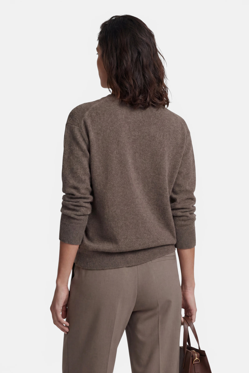 Eine Frau, von hinten gesehen, trägt einen braunen, langärmeligen Kaschmirpullover und eine dazu passende Hose. Sie hat schulterlanges, welliges braunes Haar und hält eine kleine braune Lederhandtasche an ihrer Seite, vor einem schlichten weißen Hintergrund.
