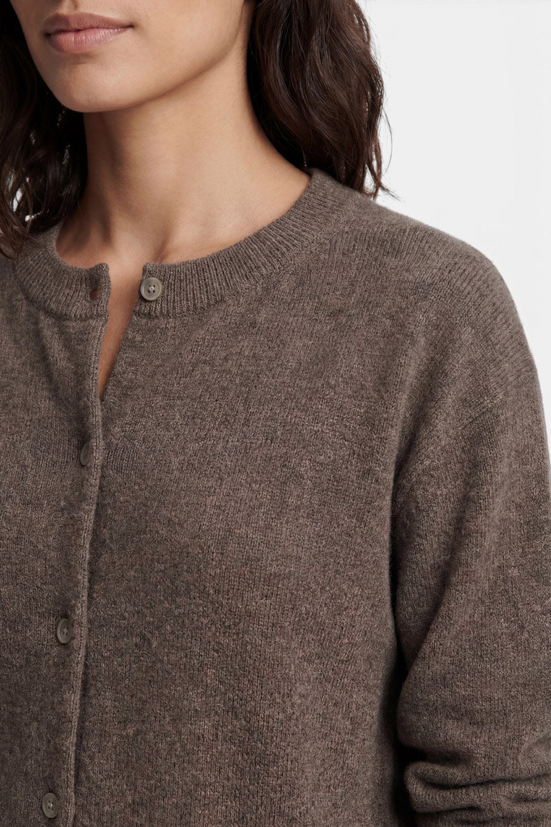 Eine Person trägt eine braune, langärmelige Kaschmir-Strickjacke mit Rundhalsausschnitt und passenden runden Knöpfen, die aus einem weichen, strukturierten Strickstoff gefertigt ist und sich von einem schlichten, hellen Hintergrund abhebt.