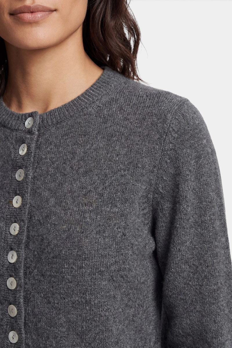 Eine Person, die eine dunkelgraue, langärmelige Kaschmir-Strickjacke trägt, die bis zum Kragen zugeknöpft ist. Der Pullover hat einen Rundhalsausschnitt mit kleinen, hellen Knöpfen, wobei der Fokus auf der Struktur des Strickstoffs und dem Oberkörper liegt.