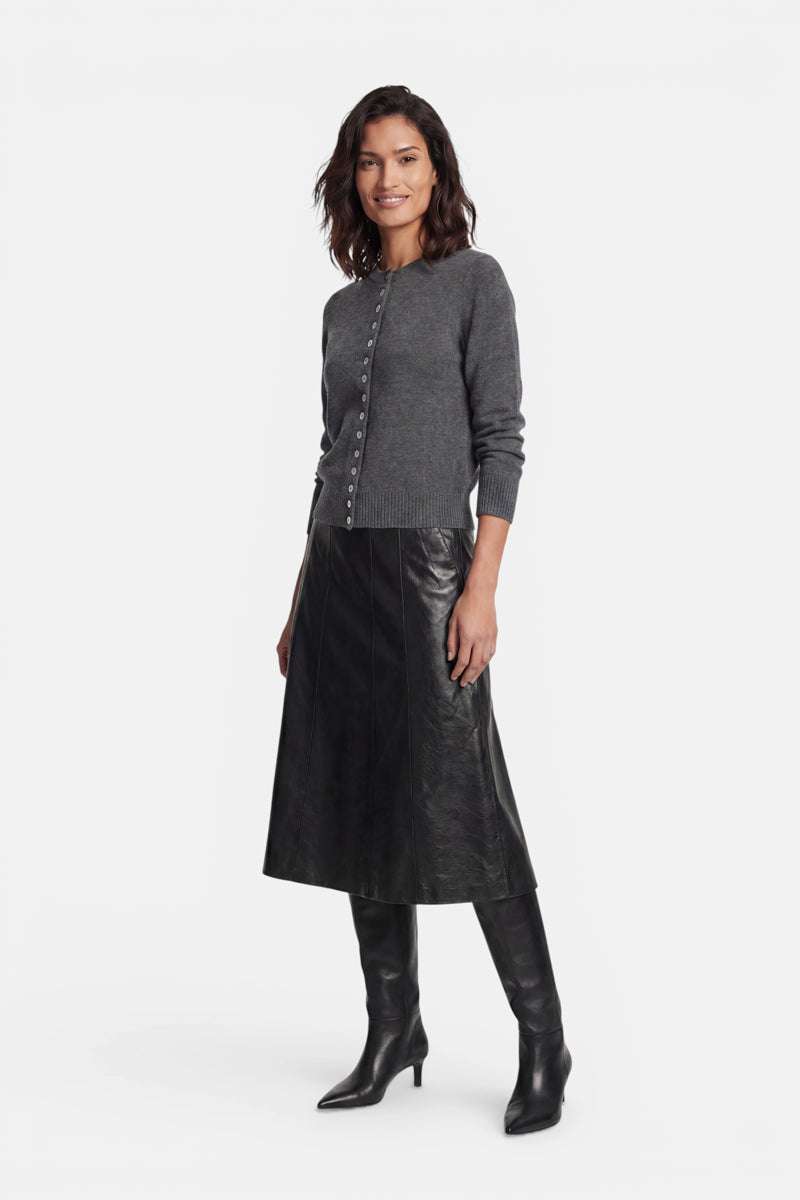 Vor einem schlichten weißen Hintergrund steht eine lächelnde Frau, die eine dunkelgraue Kaschmir-Strickjacke mit Rundhalsausschnitt, einen langen schwarzen A-Linien-Rock in Lederoptik und kniehohe schwarze Stiefel mit Absatz trägt.