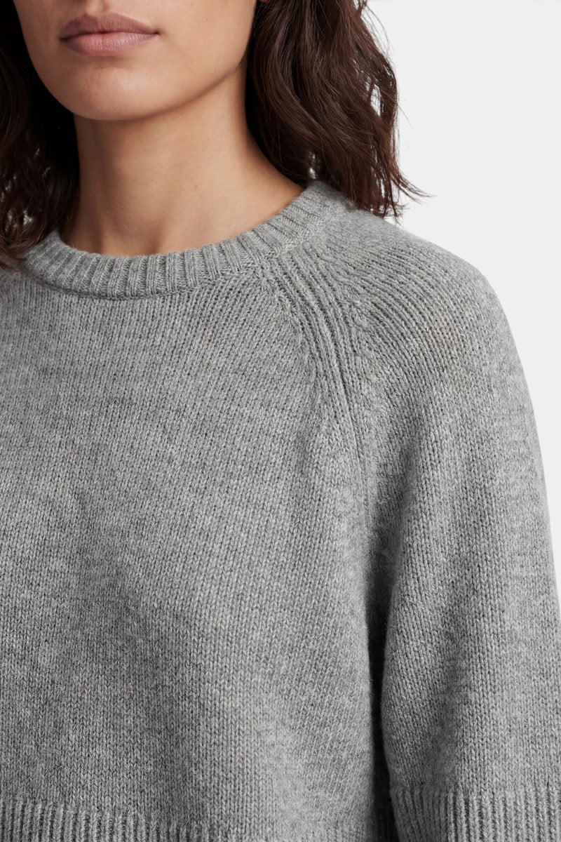 Eine Person trägt einen mittelgrauen, langärmeligen Kaschmirpullover mit rundem, geripptem Ausschnitt. Die Aufnahme konzentriert sich auf den Oberkörper und den Hals vor einem schlichten weißen Hintergrund.