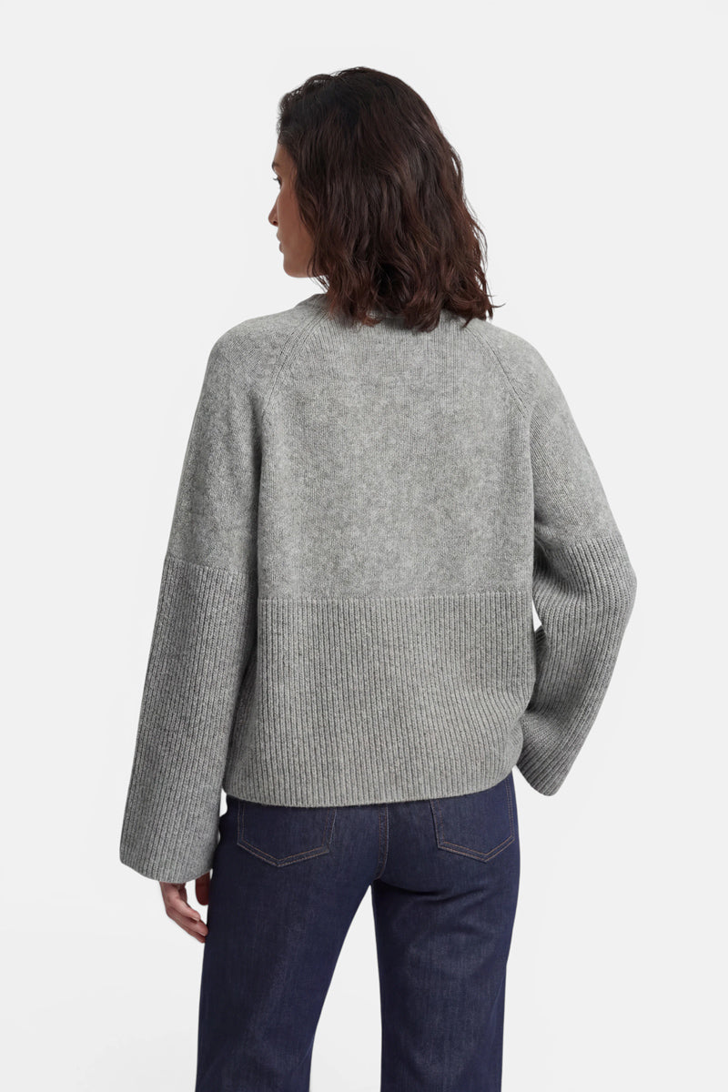 Von hinten betrachtet trägt eine Person einen grauen Kaschmirpullover in A-Linie-Schnitt, dessen unterer Teil und Ärmel mit einem breiten Rippmuster versehen sind, dazu eine dunkelblaue Jeans vor einem schlichten weißen Hintergrund.