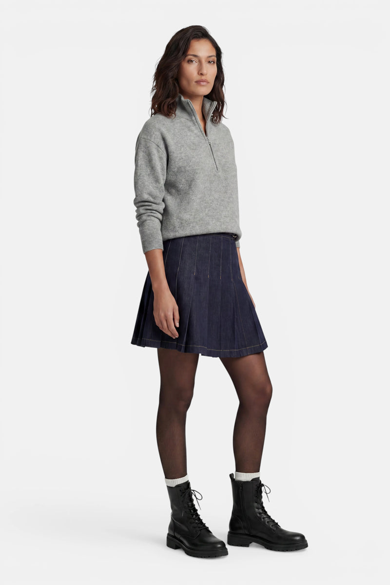 Vor einem weißen Hintergrund steht eine Frau, die einen grauen Kaschmirpullover mit Viertelreißverschluss, einen dunkelblauen Jeansrock mit Falten, eine transparente schwarze Strumpfhose, weiße Socken und schwarze Schnür-Combat-Stiefel trägt. Sie hat schulterlanges dunkles Haar und blickt in die Kamera.