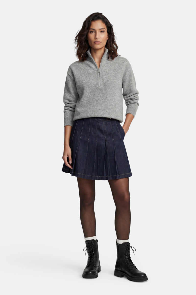 Eine Frau mit dunklem Haar trägt einen langärmeligen Kaschmirpullover mit Reißverschluss am Halsausschnitt und einen dunkelblauen, plissierten Jeansrock. Vor einem schlichten weißen Hintergrund trägt sie eine transparente schwarze Strumpfhose, weiße Socken und schwarze Schnür-Combat-Stiefel.