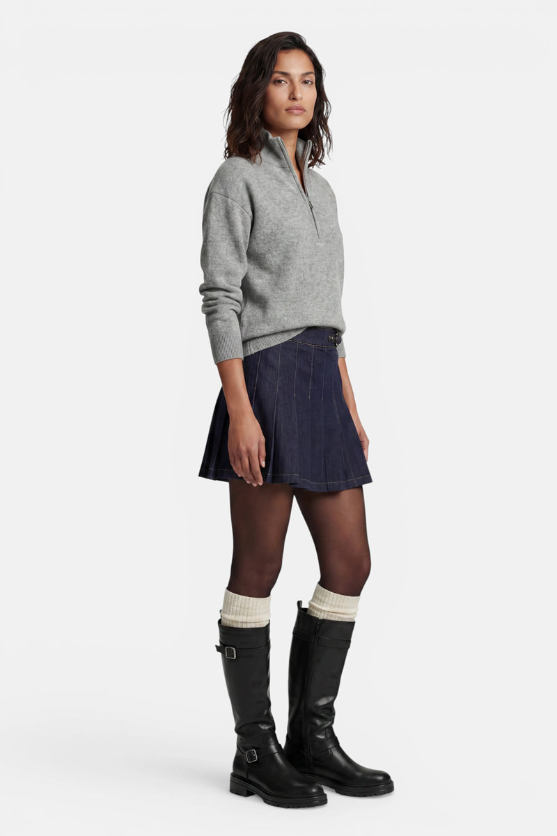 Eine Frau mit schulterlangem dunklem Haar trägt einen grauen Kaschmirpullover mit halbem Reißverschluss, der in einen dunklen Jeans-Plissee-Minirock gesteckt ist. Vor einem weißen Hintergrund trägt sie eine transparente schwarze Strumpfhose, cremefarbene gerippte Socken und kniehohe schwarze Lederstiefel.