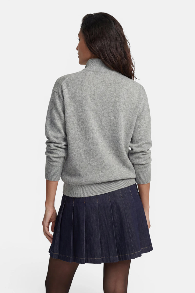 Eine Frau ist von hinten zu sehen; sie trägt einen heathergrauen Strickpullover mit Stehkragen und langen Ärmeln. Dazu trägt sie einen dunkelblauen, plissierten Jeansrock, der bis über die Knie reicht.
