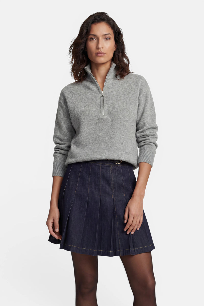 Eine Frau trägt einen grauen, langärmeligen Kaschmirpullover mit Viertelreißverschluss über einem dunklen, plissierten Jeansminirock und schwarzen, transparenten Strumpfhosen. Sie hat schulterlanges dunkles Haar und blickt vor einem schlichten weißen Hintergrund direkt in die Kamera.