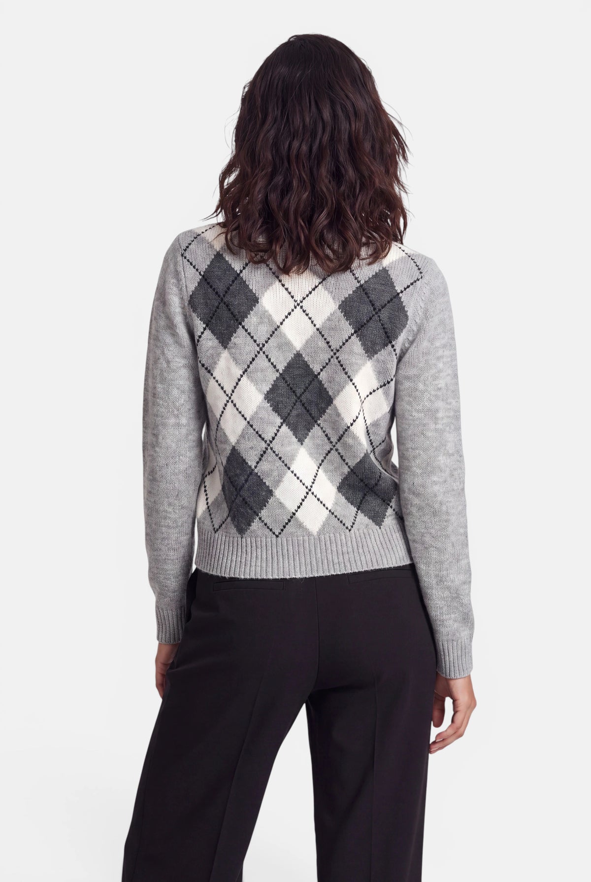Eine Frau mit schulterlangem, welligem braunem Haar, von hinten gesehen, die einen grauen, weißen und schwarzen Kaschmirpullover mit Argyle-Muster und eine schwarze Hose trägt, vor einem schlichten weißen Hintergrund.