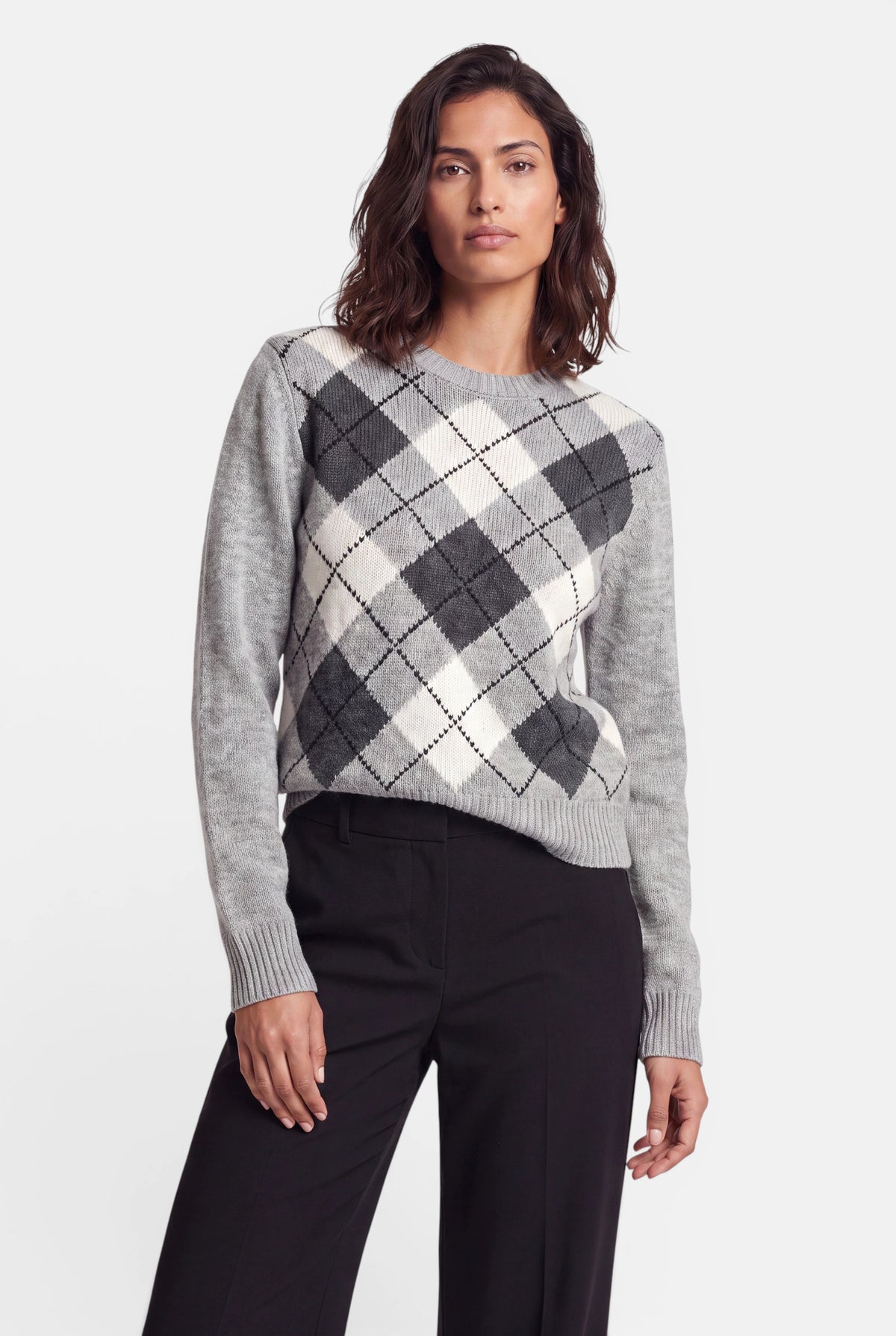 Eine Frau mit welligem braunem Haar trägt einen grauen Kaschmirpullover mit einem schwarz-weißen Argyle-Muster aus Rauten. Sie trägt eine dunkle Hose vor einem schlichten weißen Hintergrund.