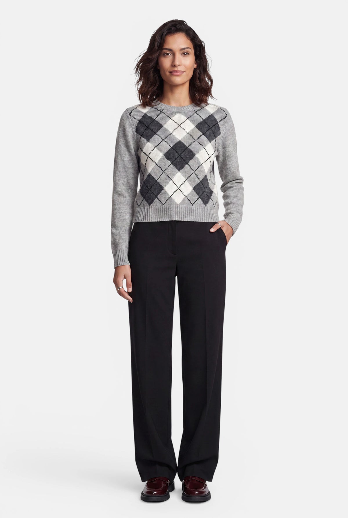 Eine Frau mit schulterlangem dunklem Haar trägt einen grau-weiß-schwarzen Argyle-Pullover mit klassischem Rundhalsausschnitt, dazu eine schwarze Hose und dunkle Slipper. Sie steht vor einem schlichten weißen Hintergrund und hat eine Hand in der Tasche.