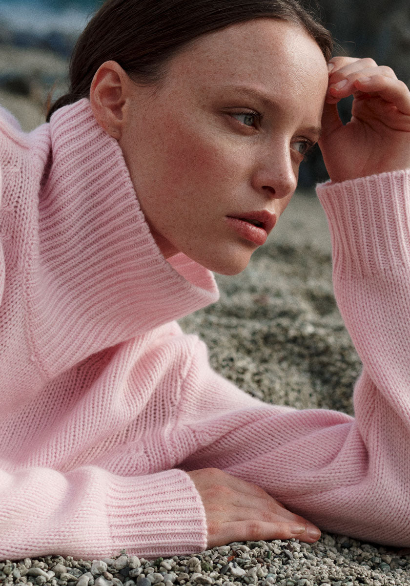 A woman with fair skin and brown hair reclines on a rocky surface, resting her head on one hand. She is wearing a soft, chunky pale pink turtleneck sweater and gazes thoughtfully into the distance, with a calm, contemplative expression.