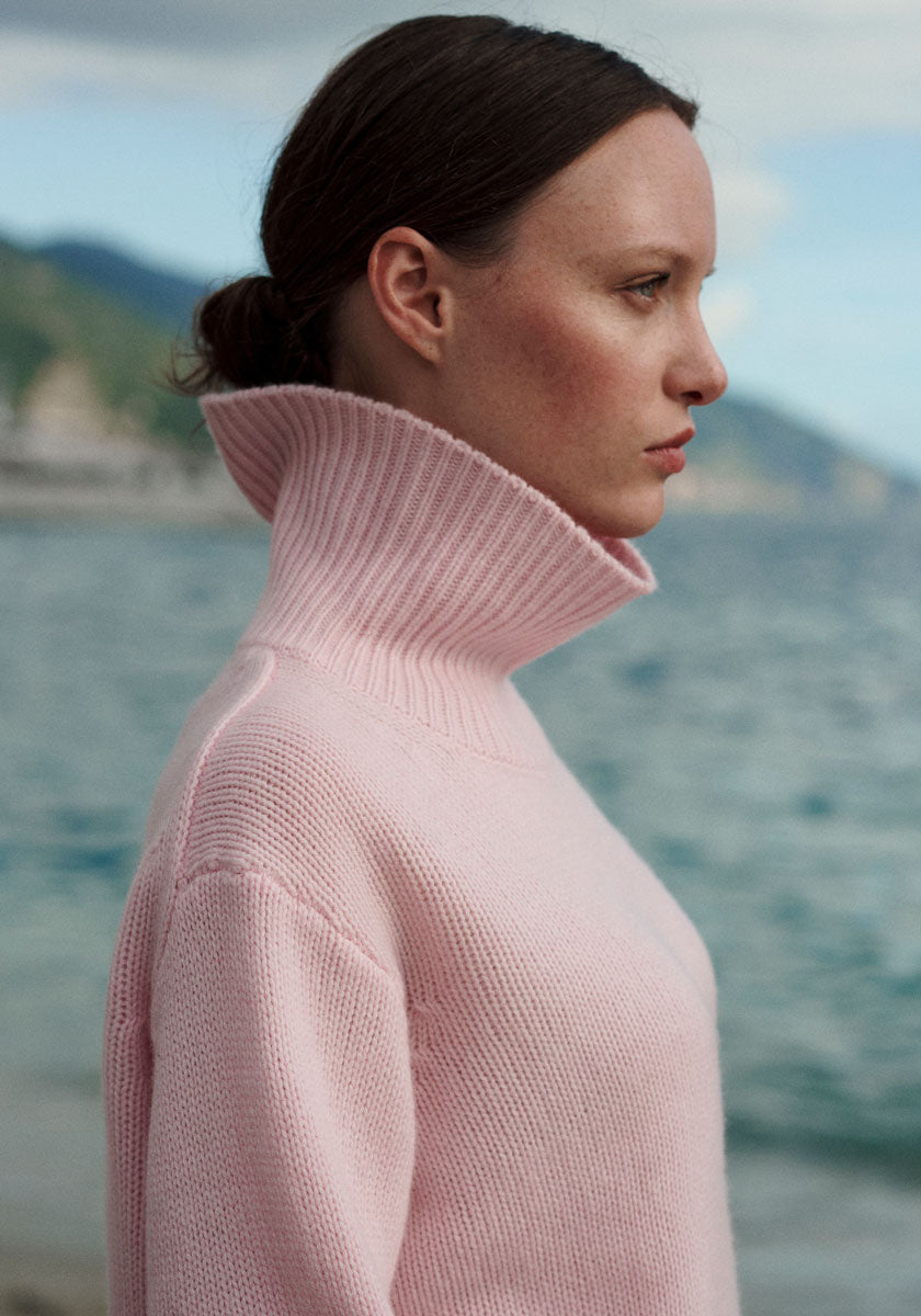 A woman with fair skin and brown hair tied back wears a light pink, high-neck sweater. She is shown in profile, standing outdoors near a calm body of water, with blurred mountains and sky in the background.
