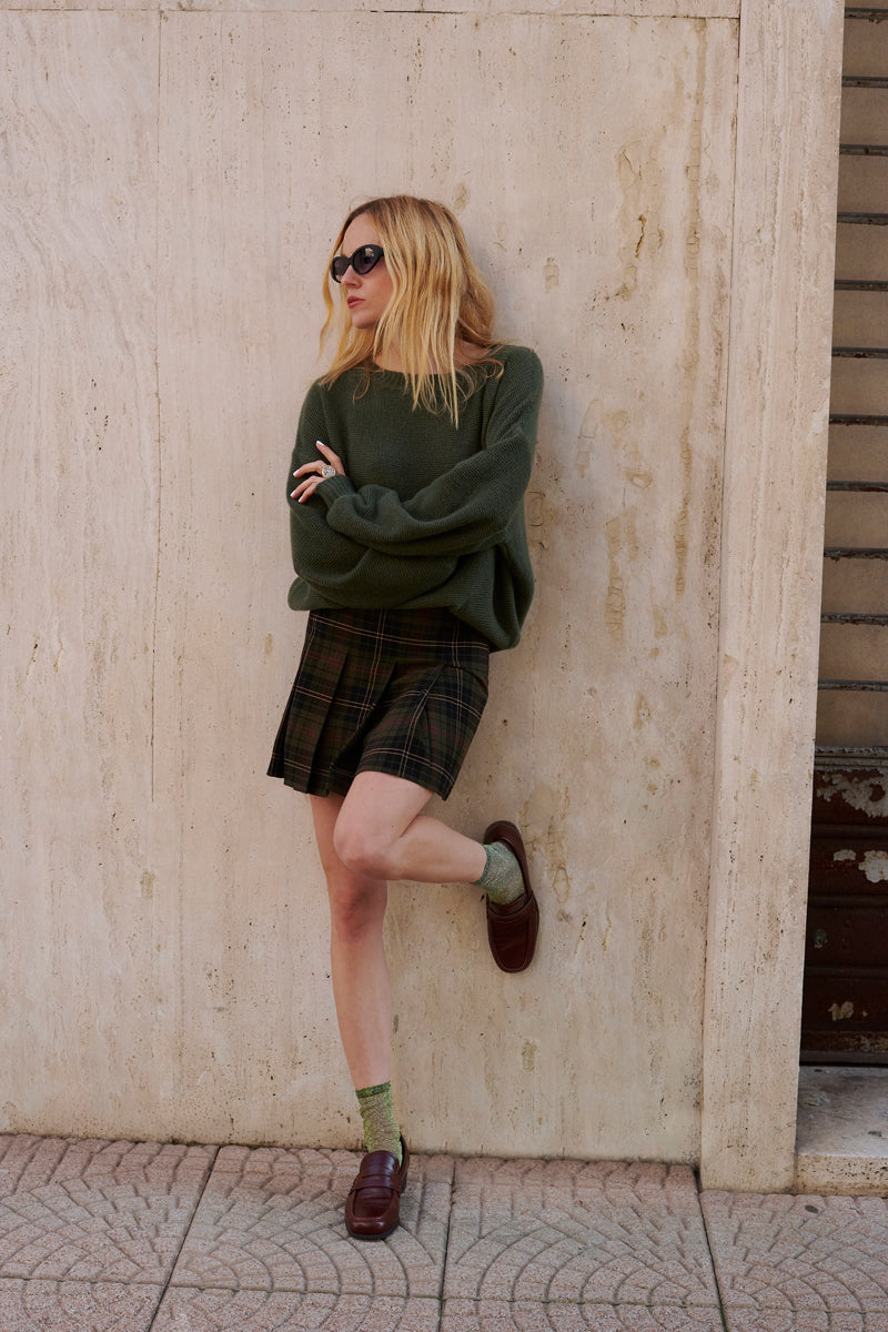 Eine Frau mit langen blonden Haaren und Sonnenbrille lehnt an einer beigen Steinmauer. Sie trägt einen übergroßen Princess Goes Hollywood Kaschmir Strickpullover mit Rundhalsausschnitt, einen dunkelkarierten Rock, grüne Socken und braune Halbschuhe. Ihre Arme sind verschränkt und ein Bein ist angewinkelt.