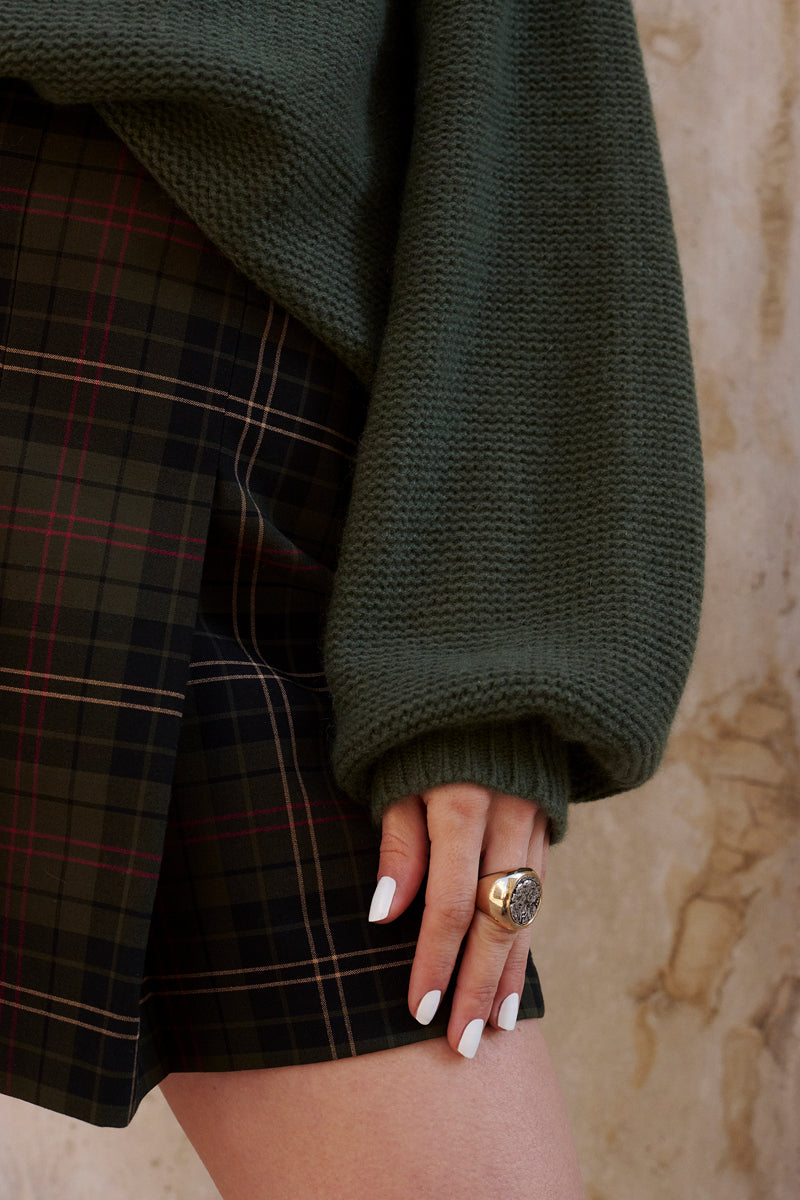 Eine Person trägt einen dunkelgrünen Kaschmir Strickpullover mit Ballonärmeln und einen dunkelgrün karierten Rock mit rosa und beigen Linien von Princess Goes Hollywood. Ihr weißer Nagellack und ihr klobiger Goldring ruhen auf ihrem Oberschenkel vor einer strukturierten beigen Wand.