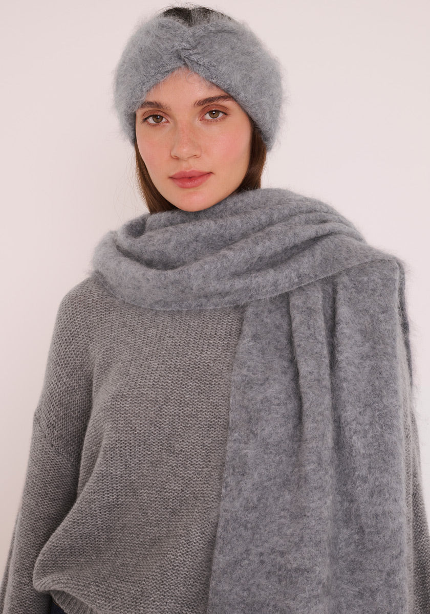 Eine Frau trägt ein weiches, flauschiges Princess Goes Hollywood Kaschmir Stirnband mit Twist-Detail und einen passenden breiten Schal. Ihr strukturierter grauer Pullover und der helle Hintergrund unterstreichen dieses gemütliche, gebürstete Kaschmir-Set.