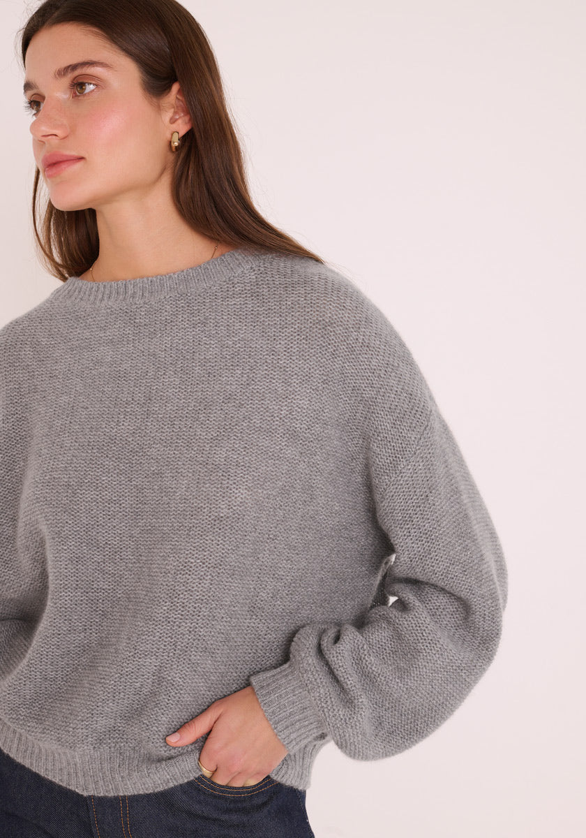 Eine Frau mit langen braunen Haaren trägt einen locker sitzenden, hellgrauen Princess Goes Hollywood Damen Pullover mit Rundhalsausschnitt und dunkle Jeans. Sie blickt mit neutralem Gesichtsausdruck leicht nach links, die rechte Hand in die Hüfte gestemmt, vor einem schlichten hellen Hintergrund.