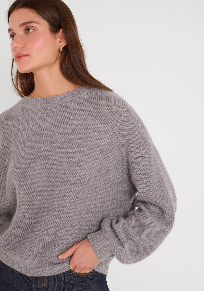 Eine Person mit langen braunen Haaren trägt einen hellgrauen Princess Goes Hollywood Kaschmir Strickpullover mit Ballonärmeln und Rundhalsausschnitt, gepaart mit einer dunklen Hose. Sie steht vor einem weißen Hintergrund und schaut neutral nach links, eine Hand in der Nähe ihrer Taille.