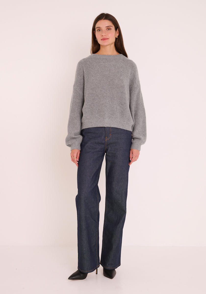 Eine Frau blickt vor einem schlichten, hellen Hintergrund nach vorne. Sie trägt einen locker sitzenden Princess Goes Hollywood Damen Pullover mit Rundhalsausschnitt, dunkelblaue High-Waisted Flared Jeans und spitze schwarze Schuhe. Ihr braunes Haar ist offen und ihre Arme sind locker an den Seiten abgelegt.