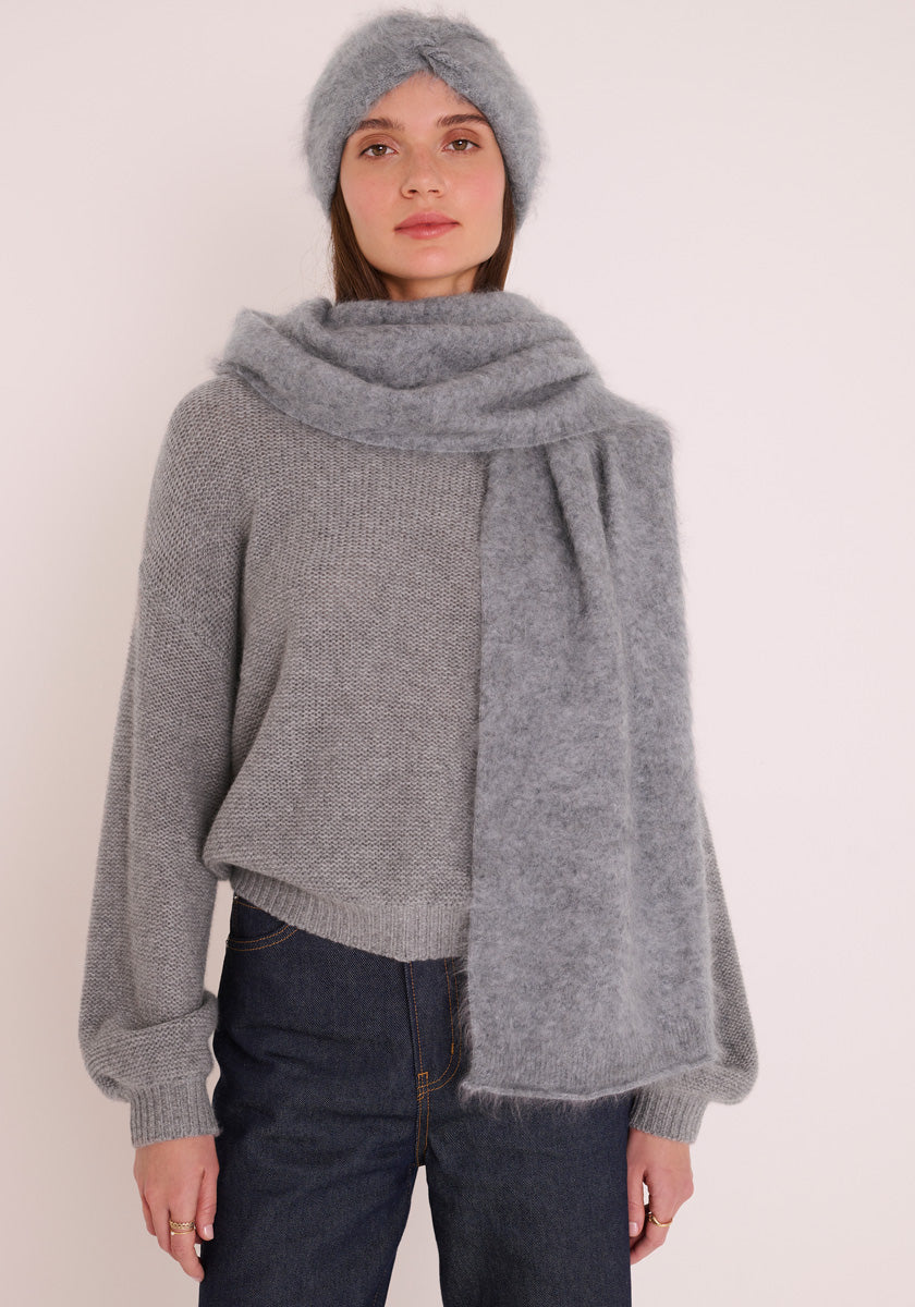 Eine Frau in einem weichen grauen Strickstirnband, einem dicken Schal, einem Princess Goes Hollywood Kaschmir Strickpullover mit Ballonärmeln und Rundhalsausschnitt und hochgezogenen dunkelblauen Jeans steht vor einem gebrochen weißen Hintergrund und blickt ruhig vor sich hin.