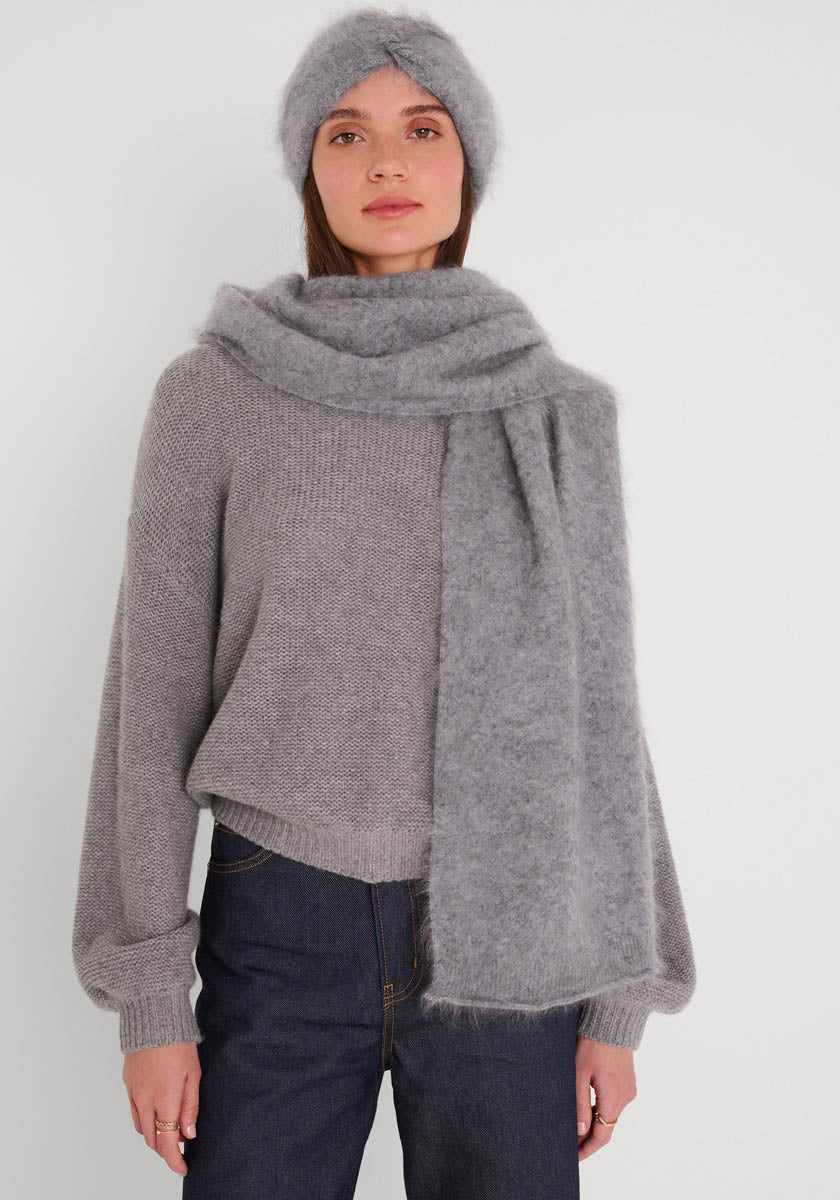 Eine Frau trägt einen weichen grauen Strickpulli mit Ballonärmeln, einen flauschigen Princess Goes Hollywood Kaschmir-Schal um den Hals und ein passendes graues Strickstirnband, gepaart mit dunkelblauen Jeans vor einem schlichten weißen Hintergrund.