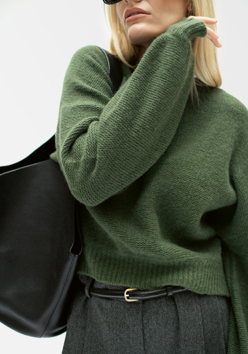 Eine Person mit langen blonden Haaren trägt einen strukturierten dunkelgrünen Princess Goes Hollywood Kaschmir Strickpullover mit Ballonärmeln, eine graue Hose mit hoher Taille und schwarzem Gürtel und trägt eine große schwarze Umhängetasche, wobei sie mit einem losen Ärmel posiert.