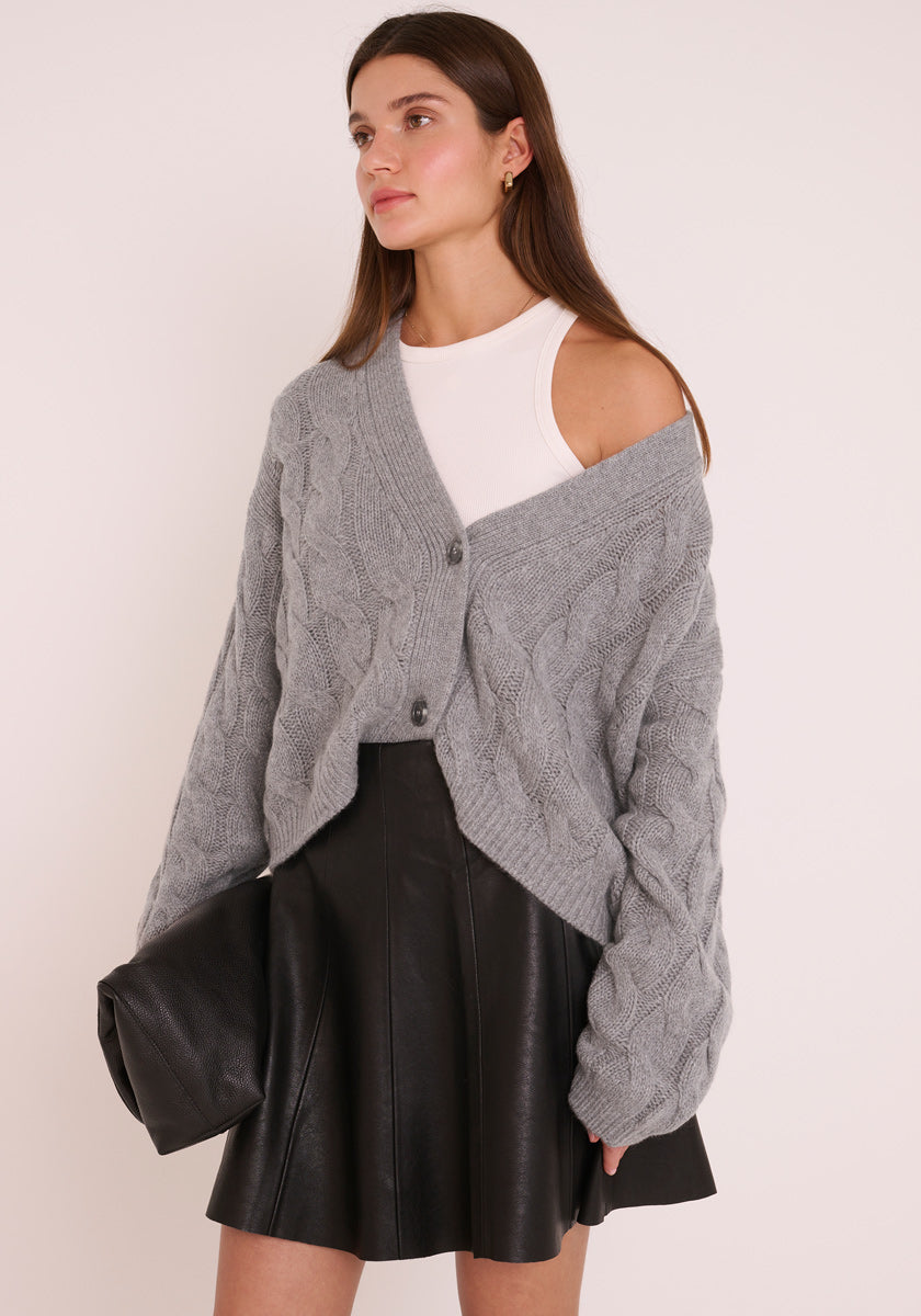 Eine Frau mit langen braunen Haaren trägt eine klobige graue Princess Goes Hollywood Zopfmuster Kaschmir Strickjacke über einer Schulter, gepaart mit einem weißen ärmellosen Top und einem schwarzen Kunstlederrock. Sie hält eine große schwarze Clutch in der Hand und schaut nach links vor einem schlichten hellen Hintergrund.