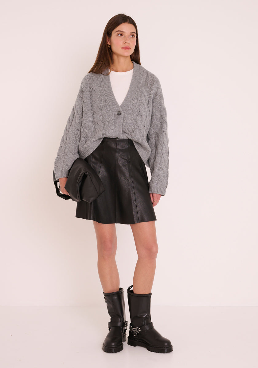 Eine Frau mit glattem braunem Haar steht vor einer weißen Wand und trägt eine strukturierte graue Princess Goes Hollywood Kaschmir Strickjacke über einem weißen Oberteil, einen schwarzen Rock in A-Linie aus Leder, schwarze wadenhohe Stiefel mit silbernen Schnallen und eine gefaltete schwarze Clutch.